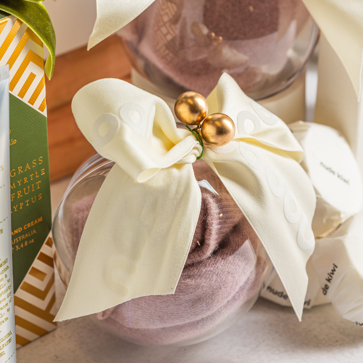 Clear Christmas bauble filled with pink Socks, tied with an ivory bow and gold decorations beside wrapped shower steamers