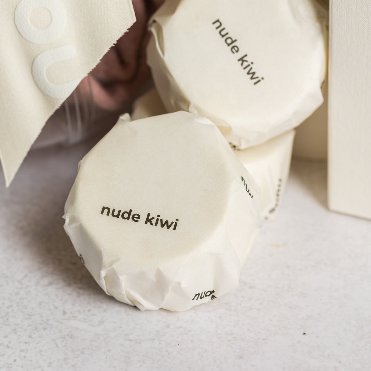 White paper-wrapped Nude Kiwi shower steamers beside a clear bauble and ivory ribbon
