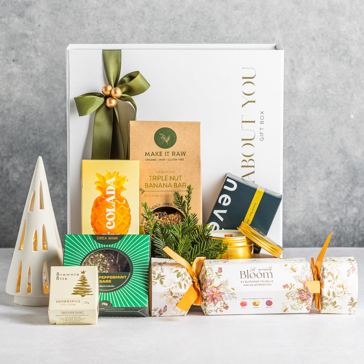Cosy Christmas gift box with a gold candle tin, floral tea cracker, sweet treats and an olive-green ribbon.