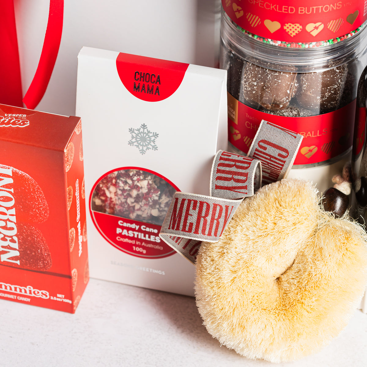 Cream bath sponge with Merry Christmas ribbon beside candy cane pastilles and jars of festive chocolates