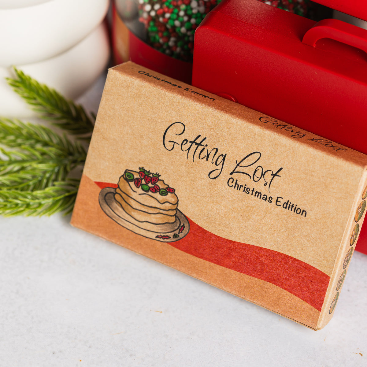 Getting Lost Christmas Edition box with a pavlova illustration, beside a red carry case and festive greenery