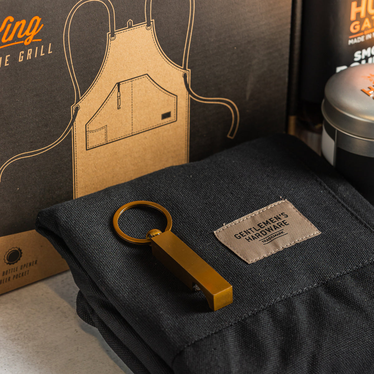 Folded black Gentlemen’s Hardware apron with a gold-coloured keyring beside its King of the Grill packaging