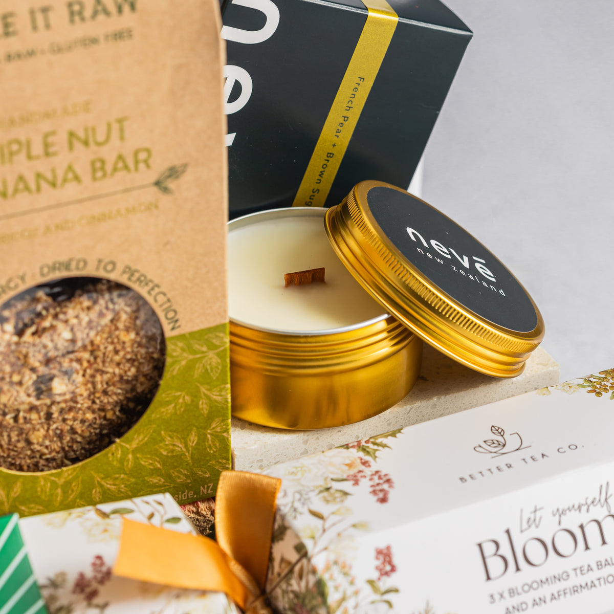 Neve New Zealand candle in an open gold tin beside a triple nut banana bar and floral tea cracker