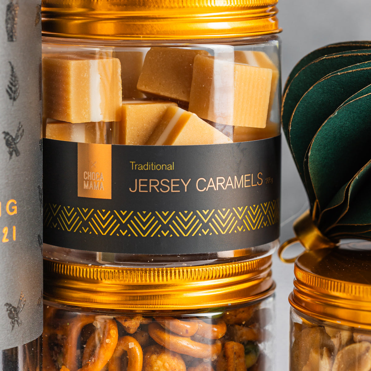 Jersey caramels in a clear gold-lidded jar above cocktail snack mix, beside a green paper Christmas bauble
