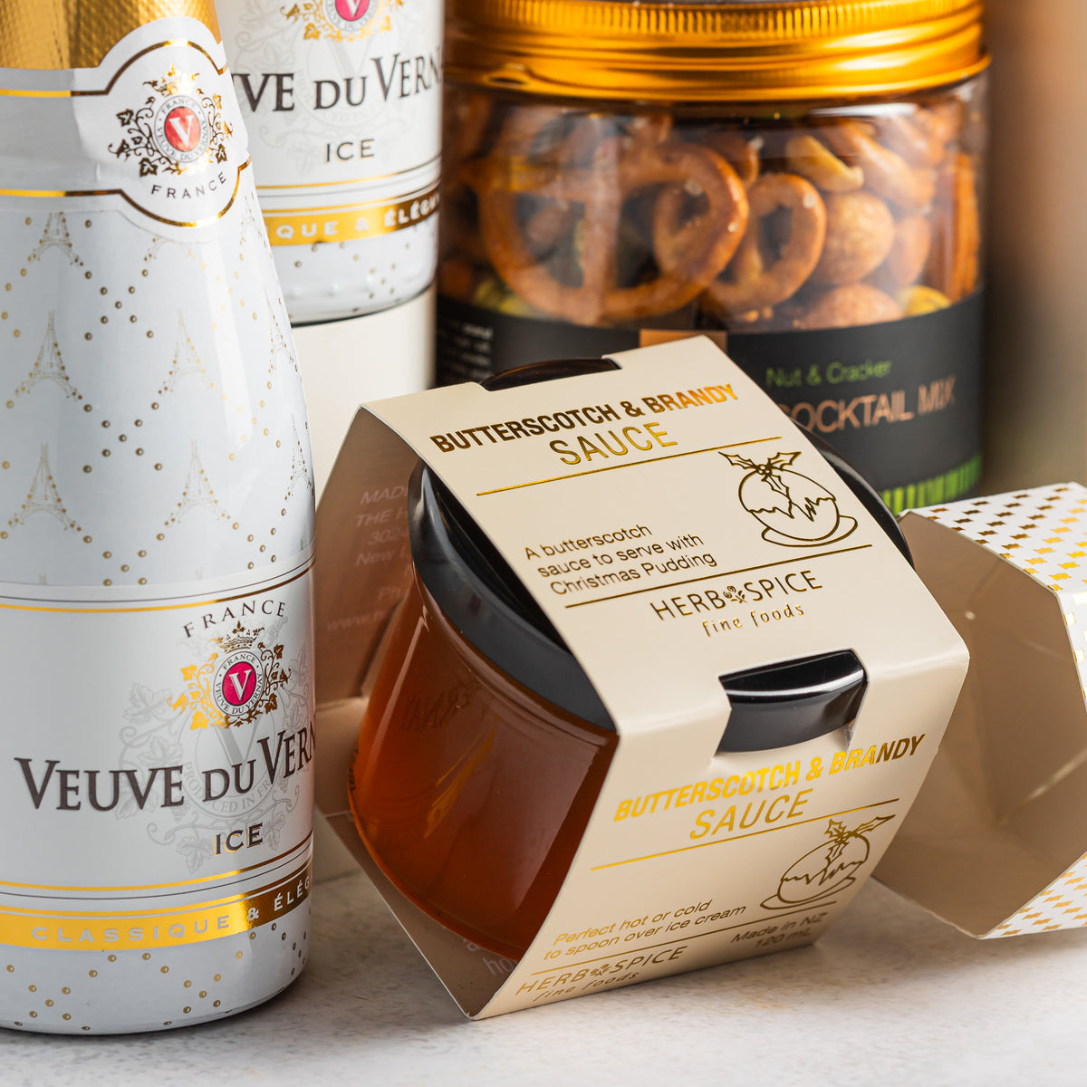Herb & Spice butterscotch and brandy sauce beside Veuve du Vernay Ice bottles and a jar of cocktail snack mix.