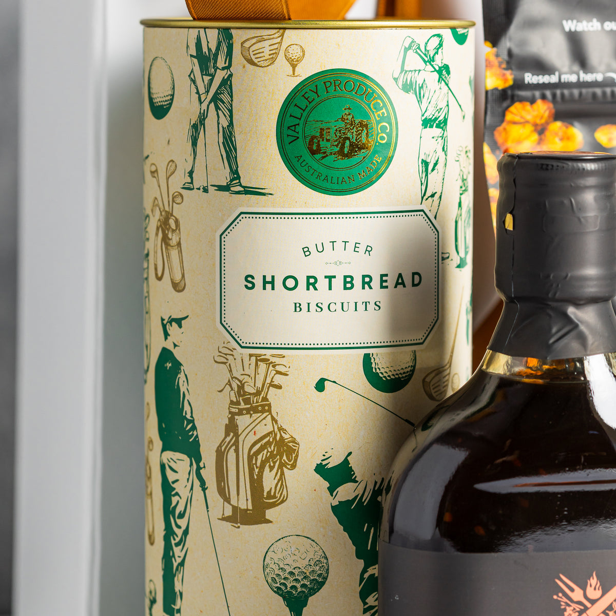 Butter shortbread biscuit tin decorated with golfers, golf clubs and balls, beside a smoky bourbon BBQ sauce bottle.